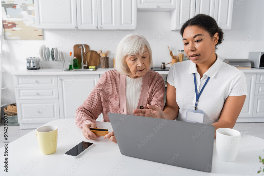 multiracial social worker using laptop while explaining senior woman with credit card how to do online shopping. multiracial social worker using laptop while explaining senior woman with credit card how to do online shopping.