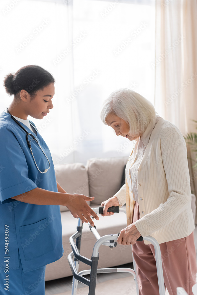 Fototapeta premium brunette multiracial nurse in blue uniform looking at senior woman with grey hair walking with help of walker at home.