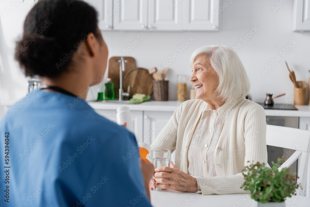 happy retired woman with grey hair looking at away near multiracial nurse on blurred foreground.