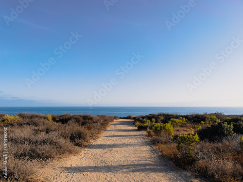 Walking path to the beach in Orange County, California on a sunny evening with low sun creating long shadows and blue ocean visible at the horizon