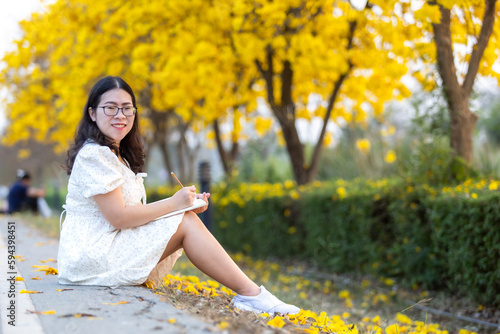 Happy Relaxing Portrait asian woman a holding diary book writing note at Yellow Tabebuia Chrysotricha flowers with the park in spring day at Evening background in Thailand.