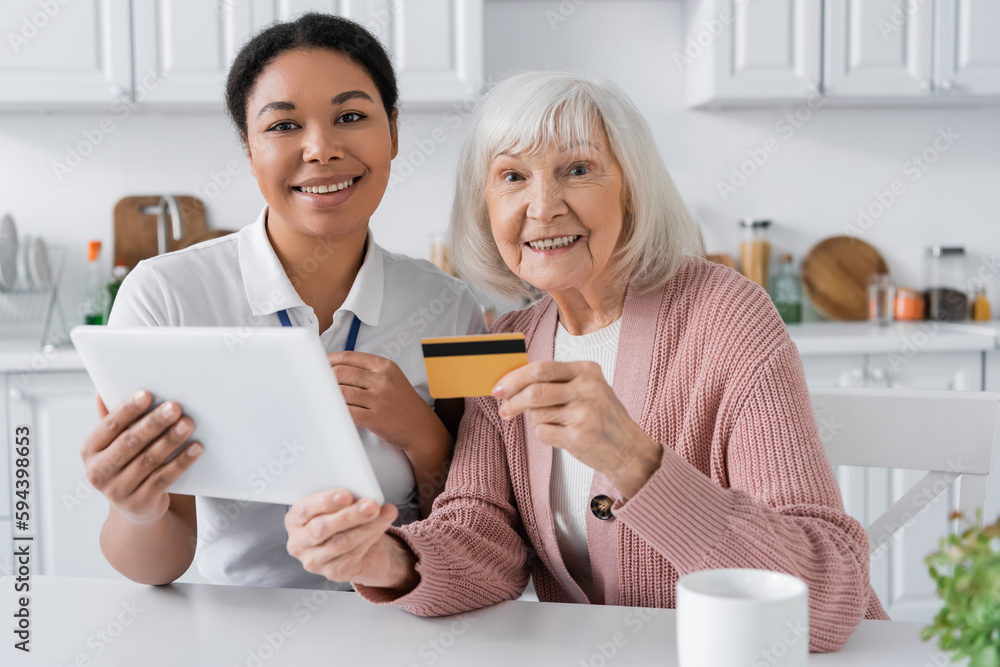 cheerful multiracial social worker holding digital tablet near positive senior woman with credit card. cheerful multiracial social worker holding digital tablet near positive senior woman with credit card.