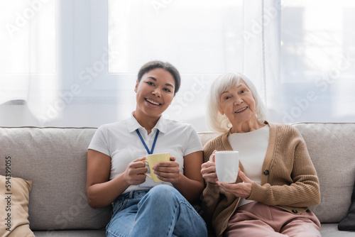 cheerful multiracial social worker having tea with senior woman in living room.