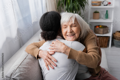 happy senior woman with closed eyes hugging curly social worker in living room. photo