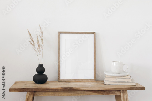 Blank vertical wooden picture frame mockup. Organic shaped black vase with dry grass on table, bench. Cup of coffee on old books. Working space, home office. Art, poster display. Boho beige interior.