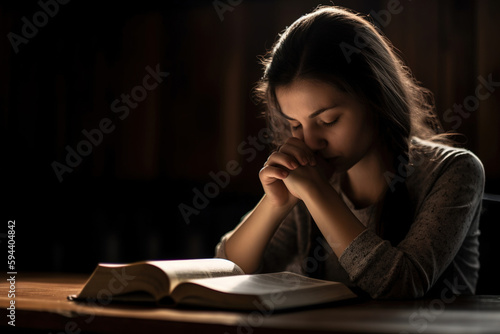 Fotografie, Tablou A young woman in a dark room reading the Bible and praying, Generation AI