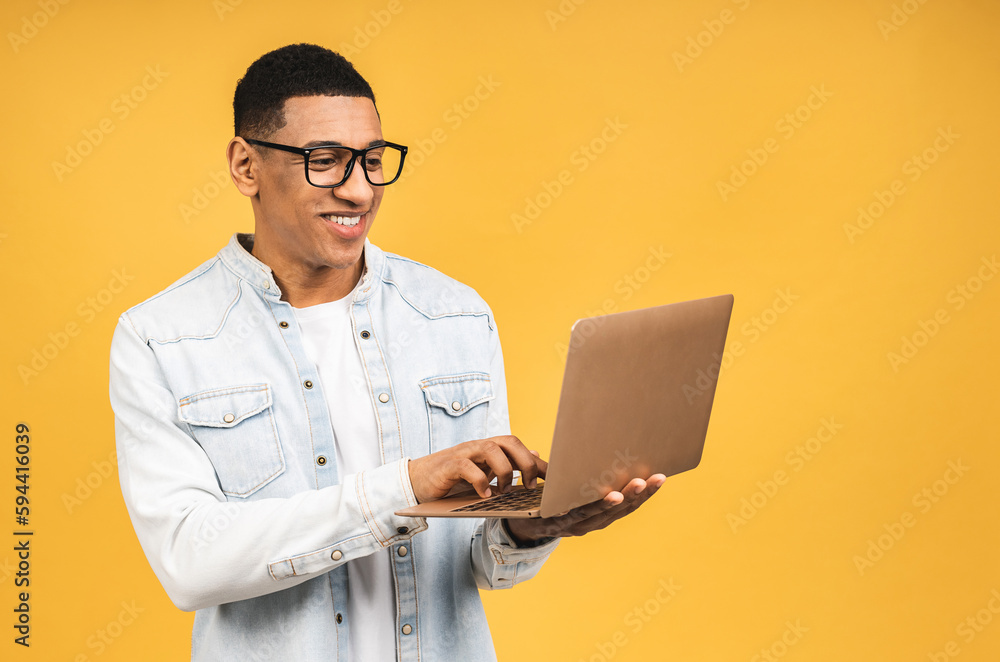 Young smiling african american man standing and using laptop computer isolated over yellow background. Young smiling african american man standing and using laptop computer isolated over yellow background.