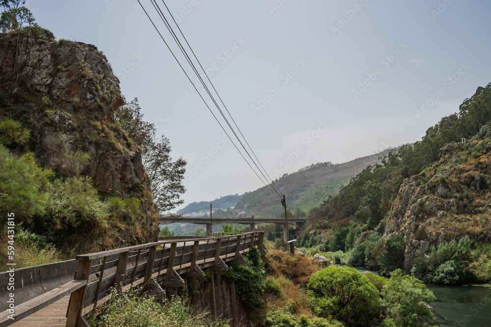 Obraz premium Wooden fence next to a hill with vegetation on the bank of the Mondego river and a bridge in the background