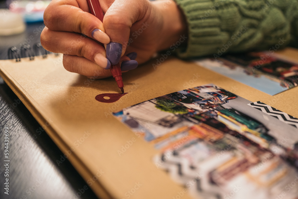 Close-up image of an unrecognizable young woman's hand filling in a red location mark with a felt-tip pen on a handmade kraft album with travel photos and washi tape.
