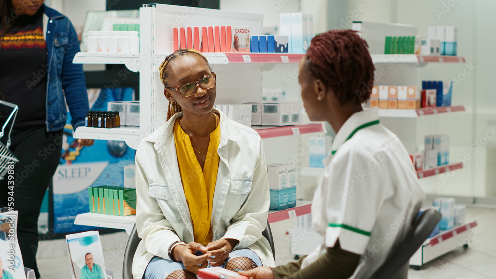 Obraz premium African american specialist showing pack of medicine to young customer, recommending pharmaceutical products in drugstore. Women talking about medicaments and vitamins in pharmacy.