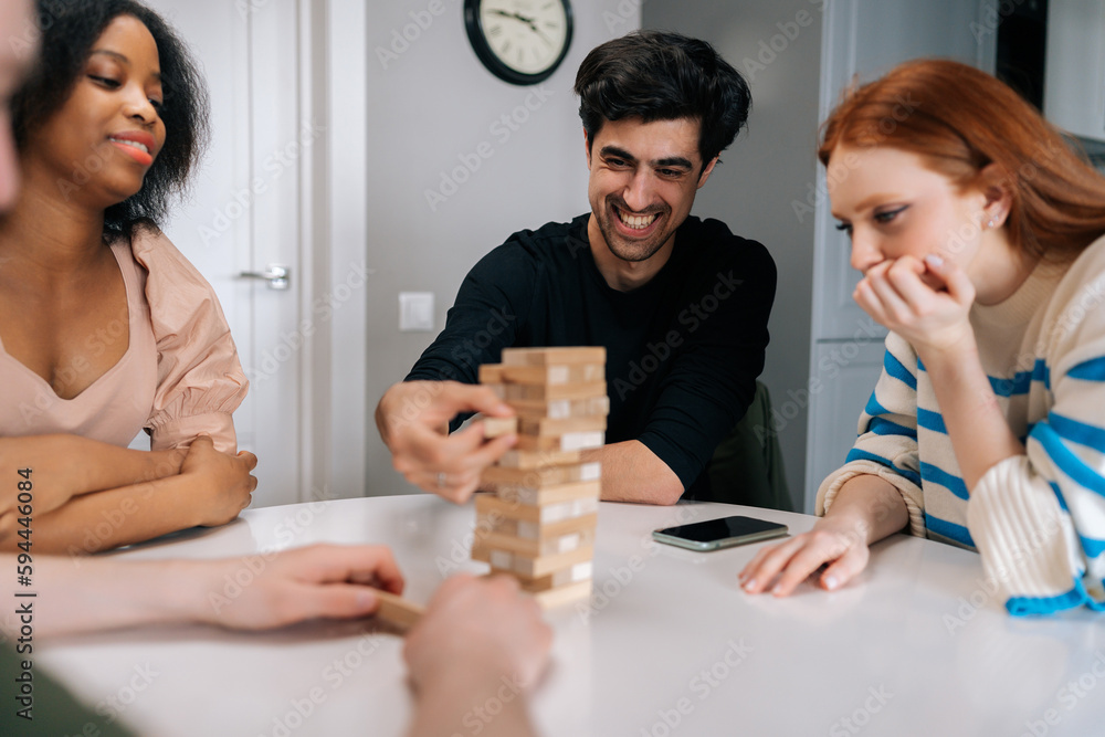 Cheerful young man carefully pulling out wood cube from wooden tower structure sitting at table. Positive happy male playing society games with multiethnic friends while enjoying fun leisure activity. Cheerful young man carefully pulling out wood cube from wooden tower structure sitting at table. Positive happy male playing society games with multiethnic friends while enjoying fun leisure activity.