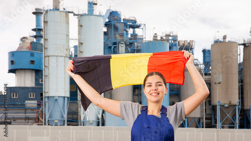 Happy young woman worker holding big flag of Belgium against background of factory photo