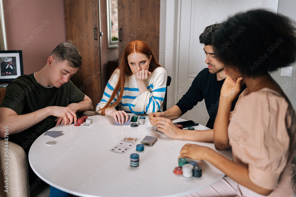 Fototapeta premium Portrait of pensive multiethnic friends having home party on weekends playing poker game in living room sitting at table. Diverse men and women satisfied with spending leisure together playing poker.
