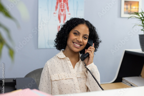 Portrait of happy biracial medical receptionist sitting at reception desk and talking on telephone