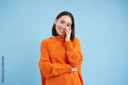 Portrait of young happy woman in orange jamper, touches her clear glowing skin, has beautiful white smile and candid emotions, blue background photo