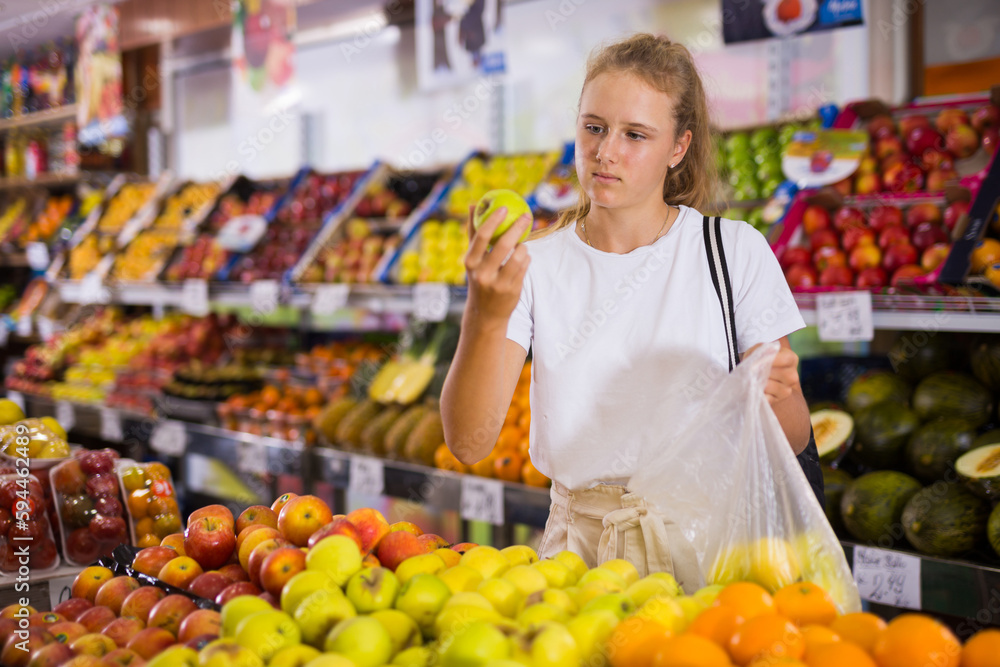 Portrait of teenage blonde girl customer buying sweet golden apples at grocery shop