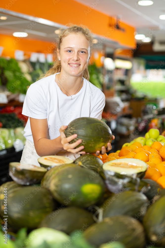 Fototapeta premium Young positive girl choosing sweet fresh melon at grocery section of supermarket