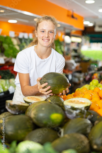 Young positive girl choosing sweet fresh melon at grocery section of supermarket