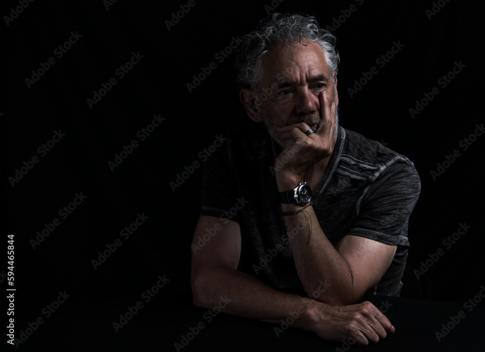 Low key color portrait of an attractive older Caucasian man with grey hair and beard. Turned to the left side with a serious or pensive expression, hand over mouth, arms visible. Room for copy.