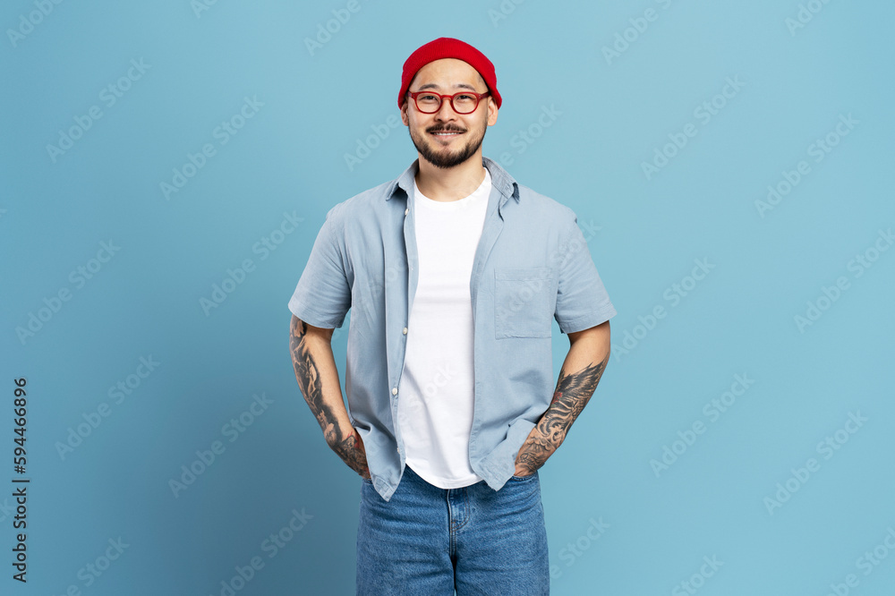 Attractive smiling asian man with stylish tattoo isolated on blue background, copy space. Happy confident Korean hipster wearing red hat, blue jeans, eyeglasses looking at camera Attractive smiling asian man with stylish tattoo isolated on blue background, copy space. Happy confident Korean hipster wearing red hat, blue jeans, eyeglasses looking at camera