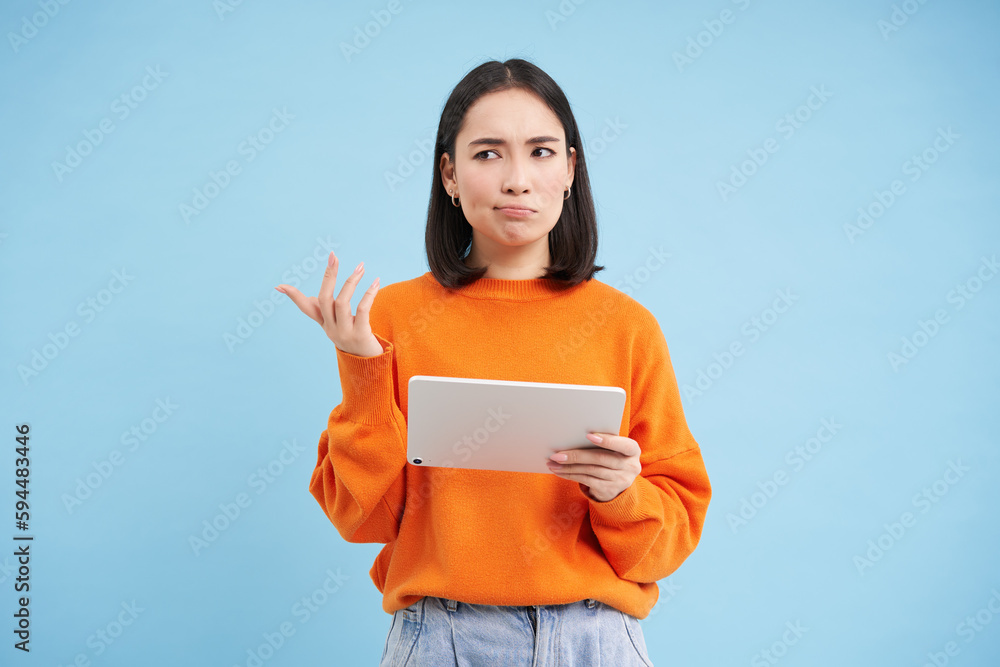 Woman with digital tablet, looks confused, cant understand smth on her gadget, stands puzzled against blue background - obrazy, fototapety, plakaty 