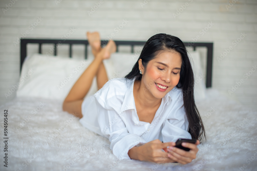 Beautiful black haired woman lays on the bed and playing her mobile phone in hotel room with smile on her face. Beautiful black haired woman lays on the bed and playing her mobile phone in hotel room with smile on her face.