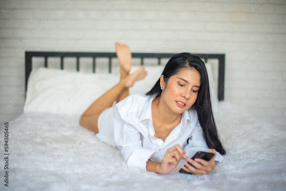 Beautiful black haired woman lays on the bed and playing her mobile phone in modern room. Beautiful black haired woman lays on the bed and playing her mobile phone in modern room.