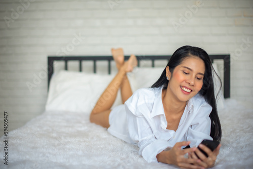 Beautiful black haired woman lays on the bed and holding her mobile phone in modern room with smile on her face. © DSM