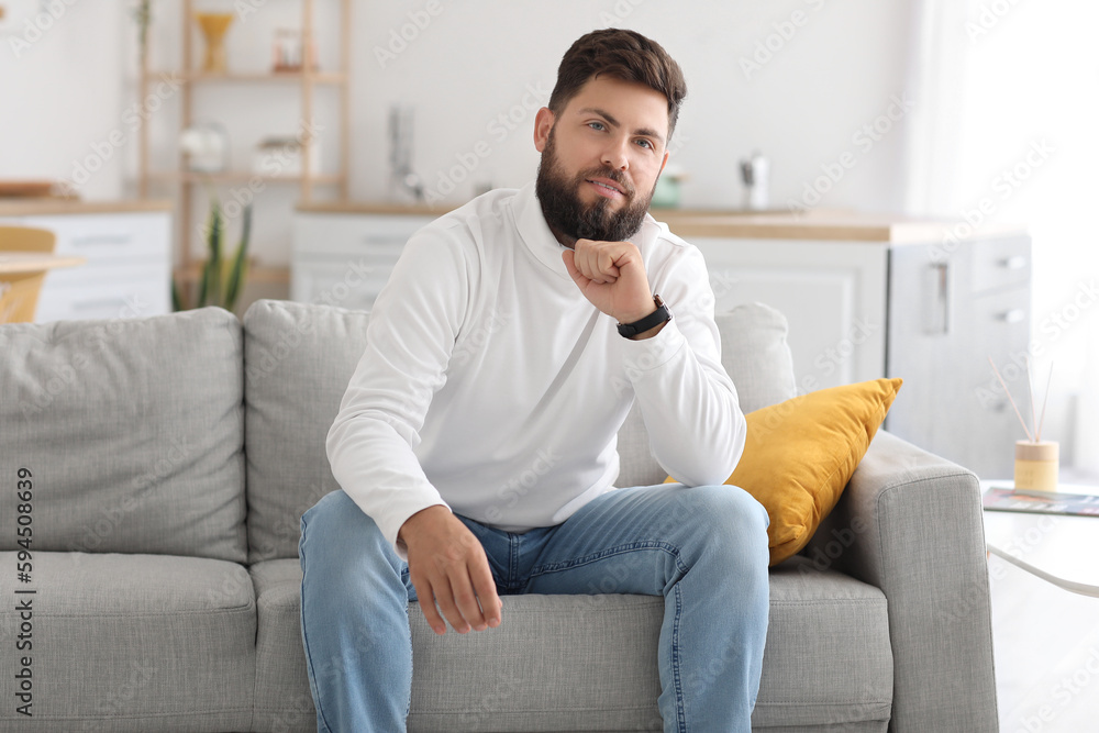 Thoughtful young bearded man sitting on sofa in kitchen