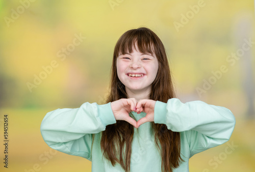 Happy young girl with Downs syndrom shows heart sign at summer park