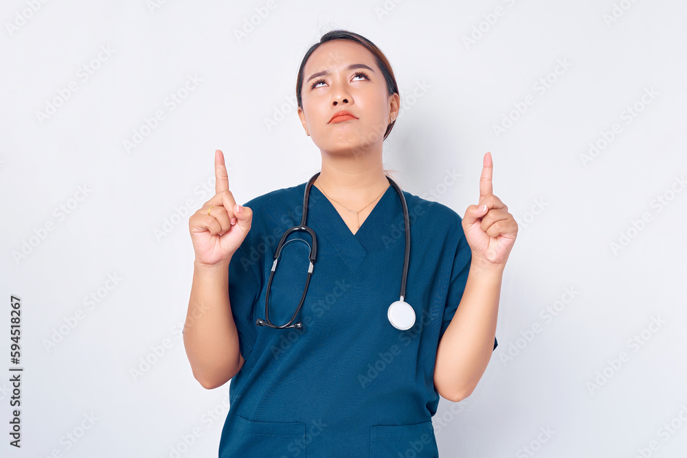 Confused young Asian female professional nurse working wearing a blue uniform pointing finger up at empty copy space isolated on white background. Healthcare medicine concept Confused young Asian female professional nurse working wearing a blue uniform pointing finger up at empty copy space isolated on white background. Healthcare medicine concept