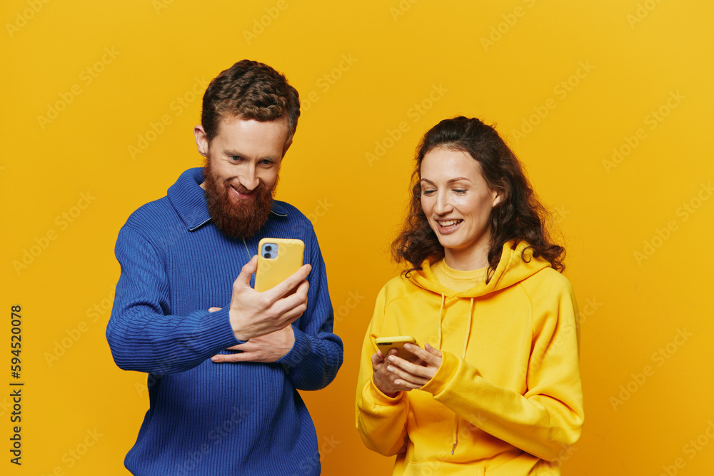 Man and woman couple smiling merrily with phone in hand social media viewing photos and videos, on yellow background, symbols signs and hand gestures, family freelancers. Man and woman couple smiling merrily with phone in hand social media viewing photos and videos, on yellow background, symbols signs and hand gestures, family freelancers.