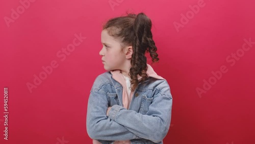 Portrait of sad unhappy little girl kid posing with crossed arms and looking sullenly to camera, feeling offended, posing isolated over pastel red color background wall in studio. Victim of bullying photo