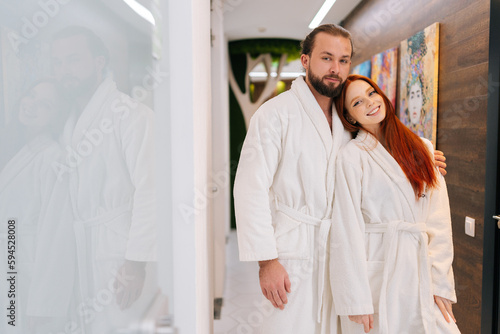 Portrait of pretty young couple in love wearing white bathrobe, standing hugging, looking at camera with smile, enjoying relaxation at spa salon. Relationship and wellness concept.