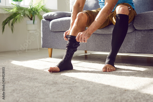 Man wears compression stockings due to varicose or recovery after thrombosis. Unrecognizable man sitting on sofa and covering tired legs with elastic medical anti varicose anti thrombotic sleeve socks photo
