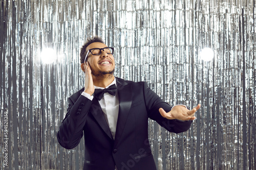 Happy funny young man in tuxedo having fun at crazy party, dancing on shiny silver foil fringe background, enjoying popular disco songs, doing hand gesture as if mixing loud music like DJ and smiling
