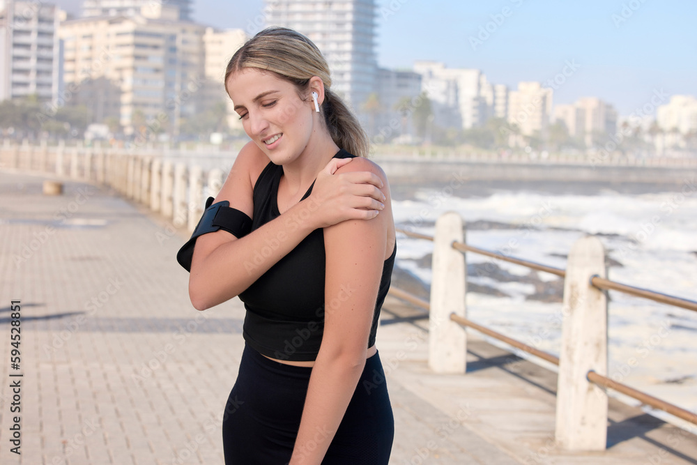 Fototapeta premium Shoulder pain, fitness woman and music of a person running by the sea with a arm injury. Workout, sports and training accident of a female runner on a exercise run and muscle strain on arms outdoor