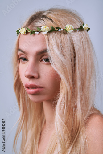 Close-up shot of a young blonde woman with a handmade wreath decorated with pink and purple flowers. A beautiful girl with long hair with a wreath on her head is on a light background. Front view.