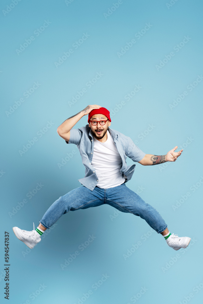 Handsome excited asian man jumping high, having fun isolated on blue background. Portrait of emotional Korean dancer wearing red hat, stylish eyeglasses, white shoes. Store, shopping, sales concept Handsome excited asian man jumping high, having fun isolated on blue background. Portrait of emotional Korean dancer wearing red hat, stylish eyeglasses, white shoes. Store, shopping, sales concept