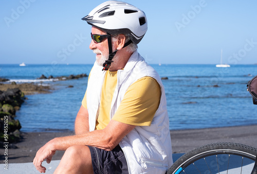 Carefree senior cyclist man with sport helmet sitting at the beach close to electric bike looking at horizon. Elderly male enjoying healthy lifestyle in retirement