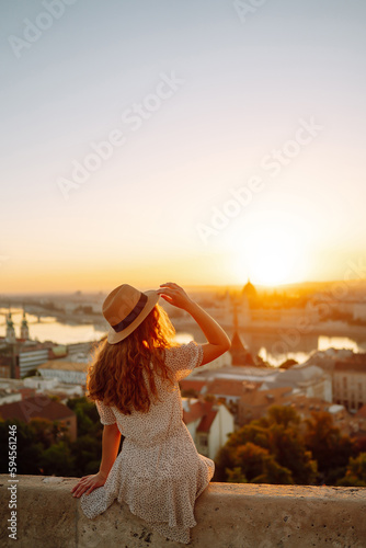 Smiling female tourist in a hat admires the landscape of the city at dawn. Euro-trip. Travel, tourism and active lifestyle concept.