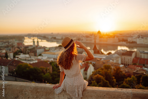 Fényképezés Smiling female tourist in a hat admires the landscape of the city at dawn