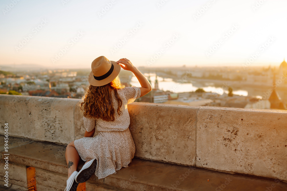 Smiling female tourist in a hat admires the landscape of the city at dawn. Euro-trip. Travel, tourism and active lifestyle concept.