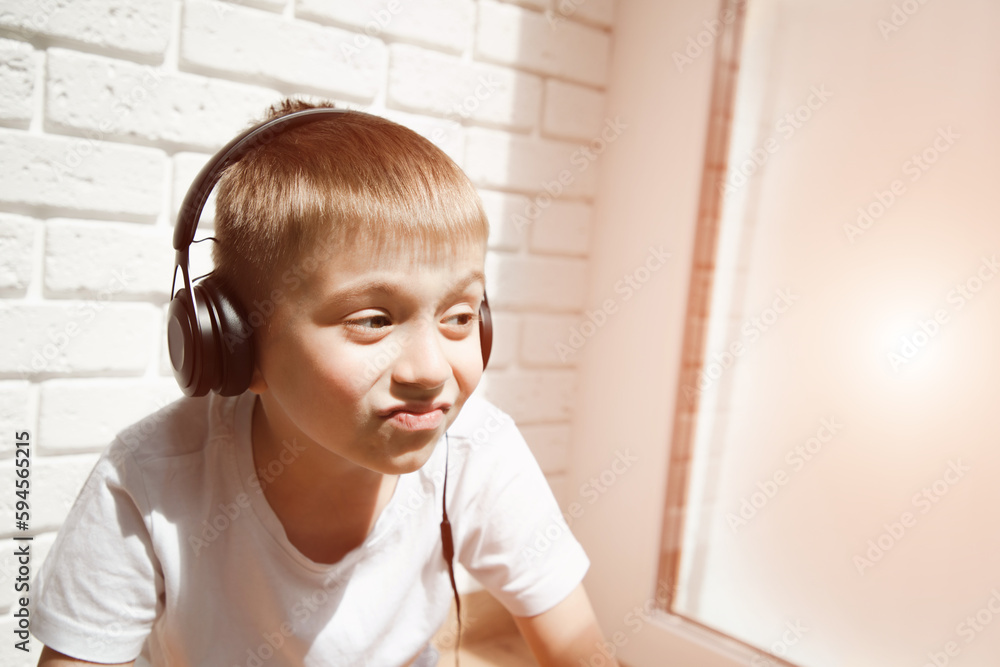 A boy of 6-7 years old in a white T-shirt listens to music in black headphones and looks out the window A boy of 6-7 years old in a white T-shirt listens to music in black headphones and looks out the window