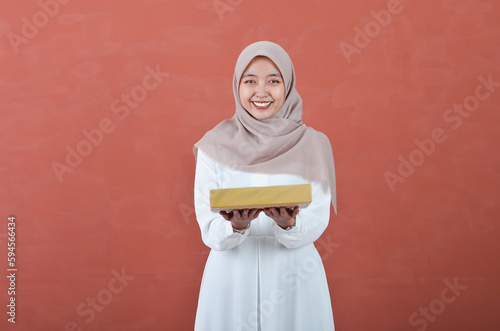 Beautiful Asian woman in white gamis and hijab smiling cheerfully bring a giift box isolated in brown background photo