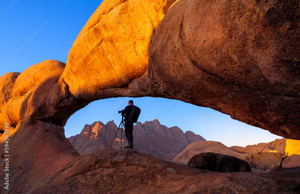 Nature photographer takes photo on his camera with tripod in a stone arch of the rock on Spitzkoppe, Namibia. Calm landscape at sunrise with silhouette of tourist in a mountain valley on Spitzkoppe.