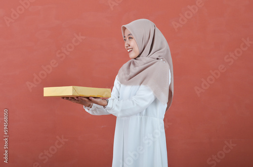 Beautiful Asian woman in white gamis and hijab smiling cheerfully bring a giift box isolated in brown background photo