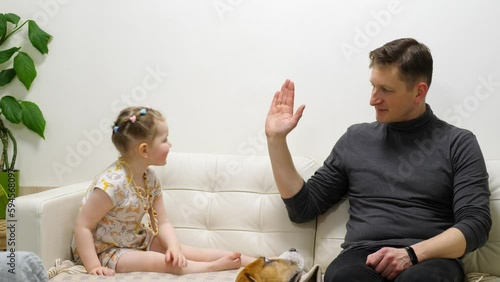 Father raise right hand and ask little daughter to give five, they slap hands and then repeat. Two people sit on sofa at living room, agreed or celebrate something, dog sit on the floor