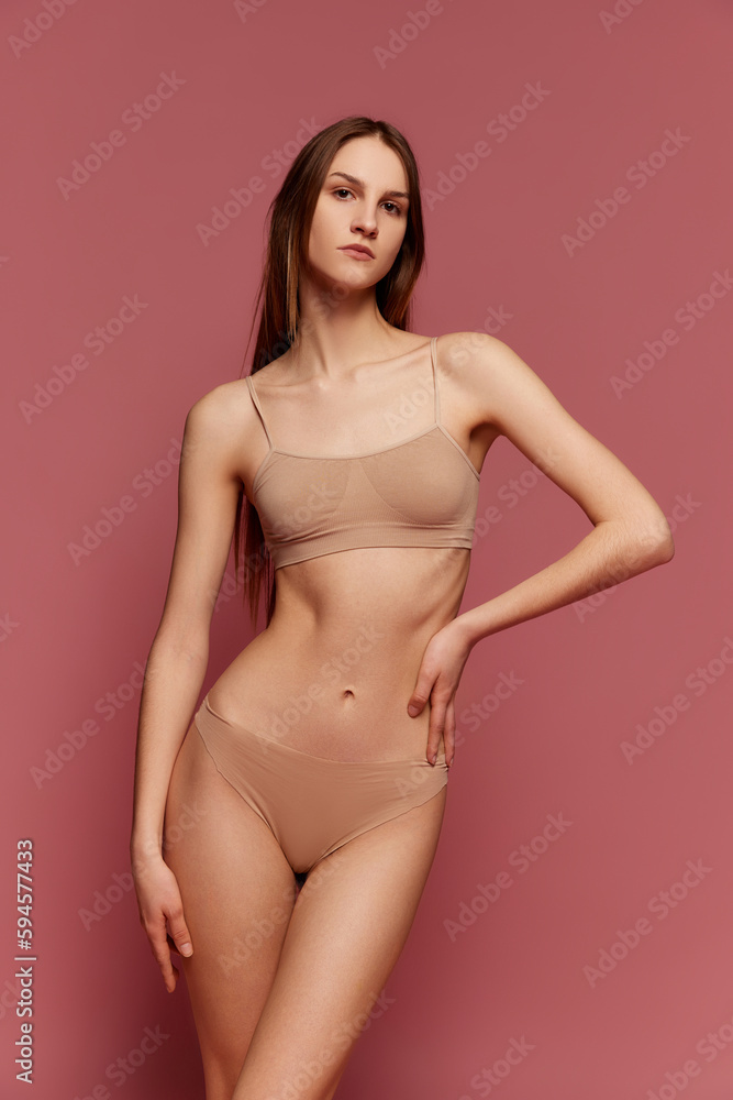 Portrait of young girl with serious face posing in beige underwear against pink studio background. Slim, muscular body shape. Concept of beauty, body care, fitness, sport, health, figure Portrait of young girl with serious face posing in beige underwear against pink studio background. Slim, muscular body shape. Concept of beauty, body care, fitness, sport, health, figure