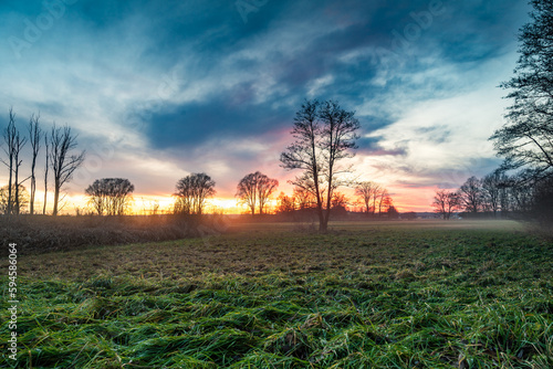 Beautiful sunset with dramatic sky and clouds on a meadow in winter with bare trees in the background